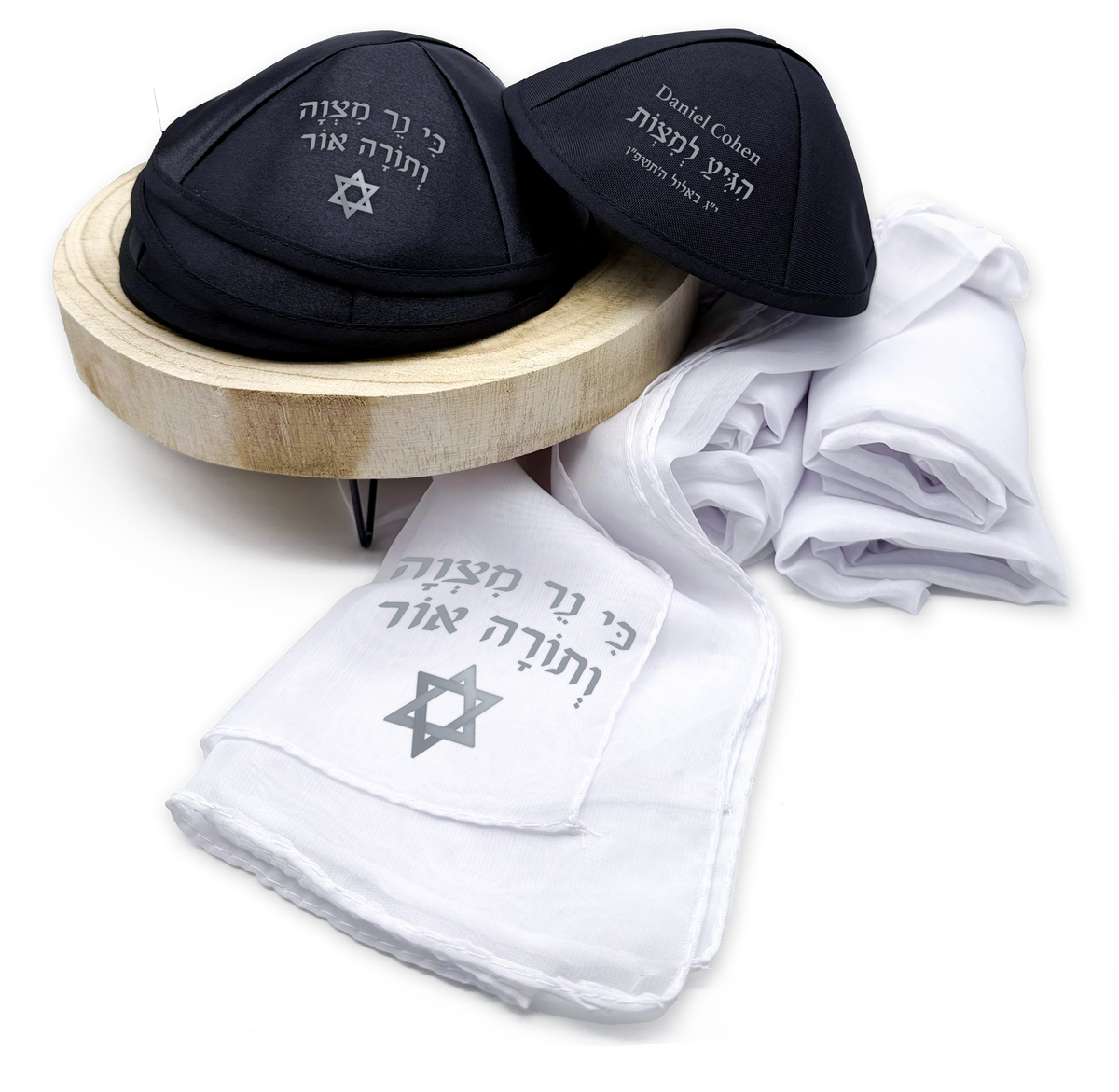 Black satin Bar Mitzvah kippahs with personalized Hebrew and English artwork beside matching white shawls